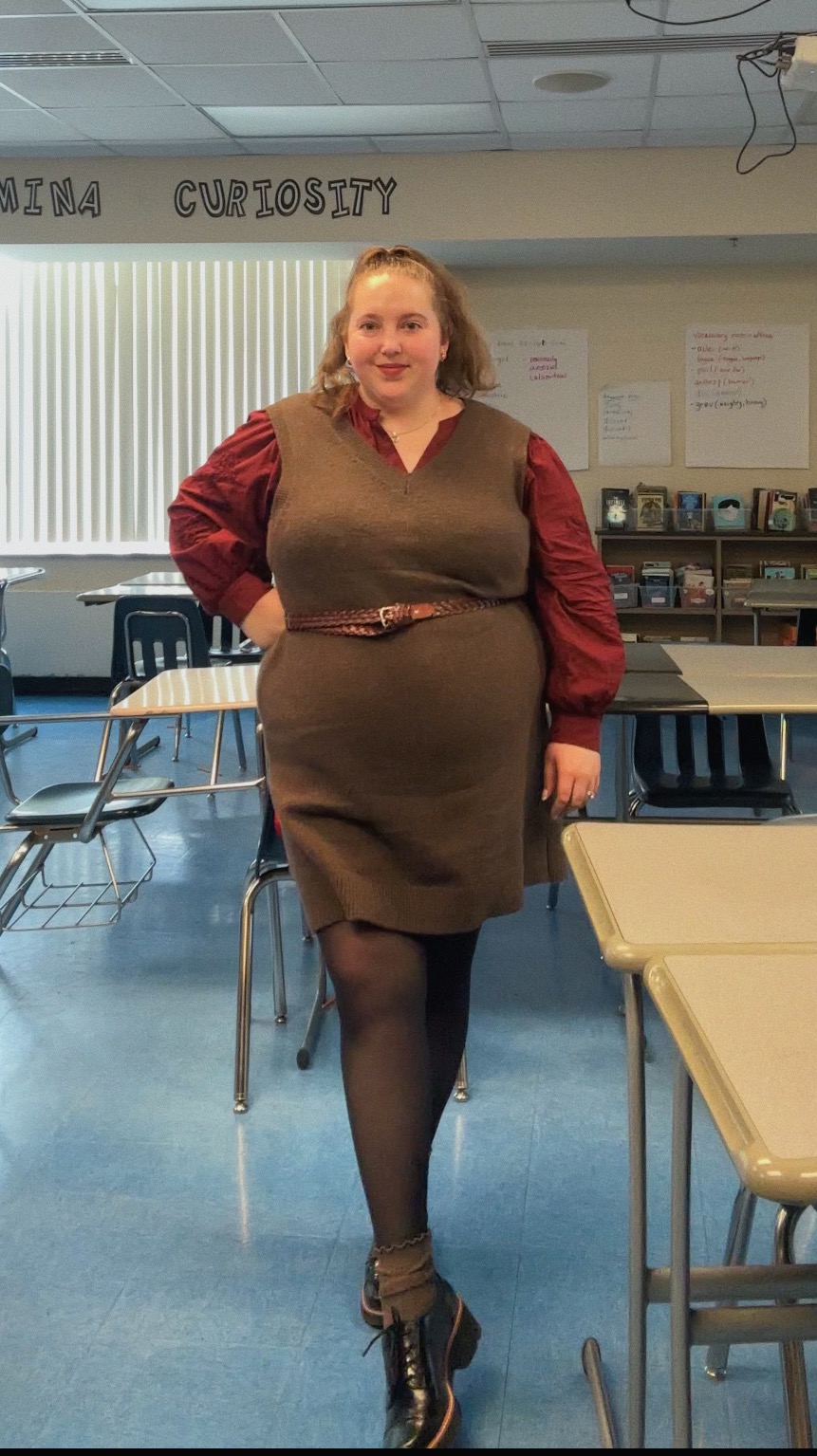 Julia Ludovici, a white woman with long brown hair up in a ponytail, wears a brown sleeveless sweater dress with a red blouse underneath. The dress is belted at the waist with a reddish-brown leather belt. She wears sheer black tights, brown ankle socks, and black leather loafers. She stands in a classroom with a blue linoleum floor surrounded by desks. Julia is a substitute teacher and provides literacy tutoring services.